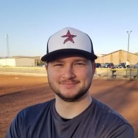 Smiling person in a navy shirt and white cap standing on a dirt field at sunset