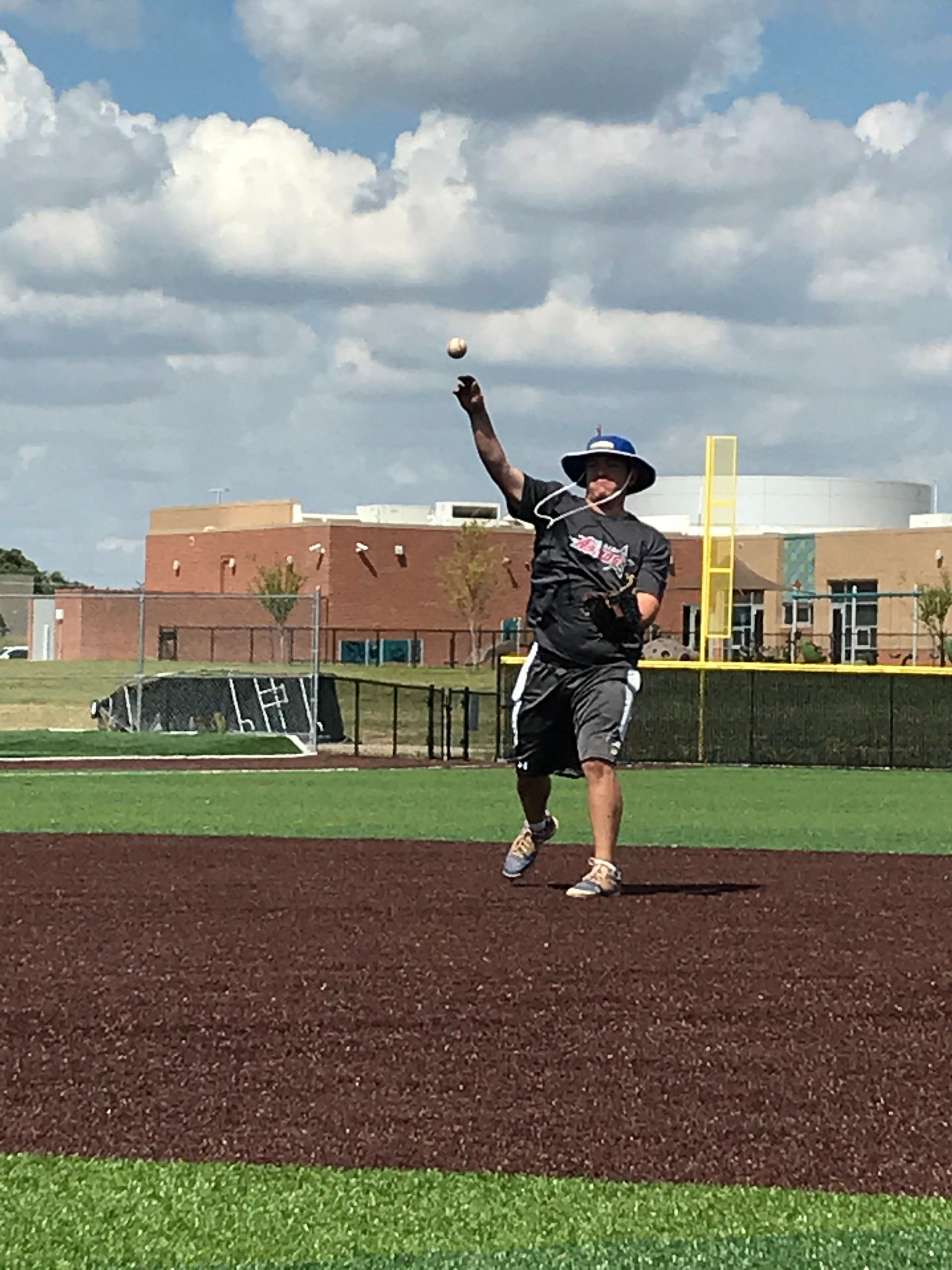 Baseball pitcher in black uniform throws a ball on a field under a cloudy sky.