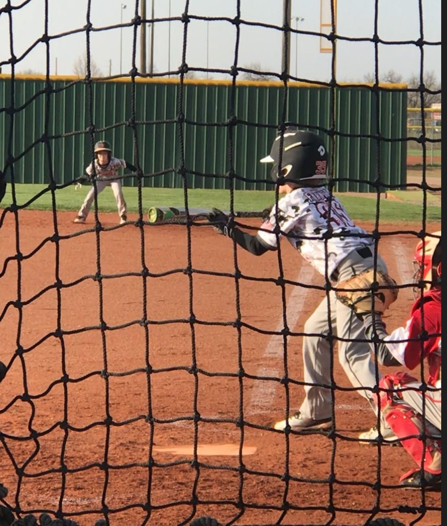 Youth baseball batter at the plate, catcher behind him, and pitcher on the mound behind the net.