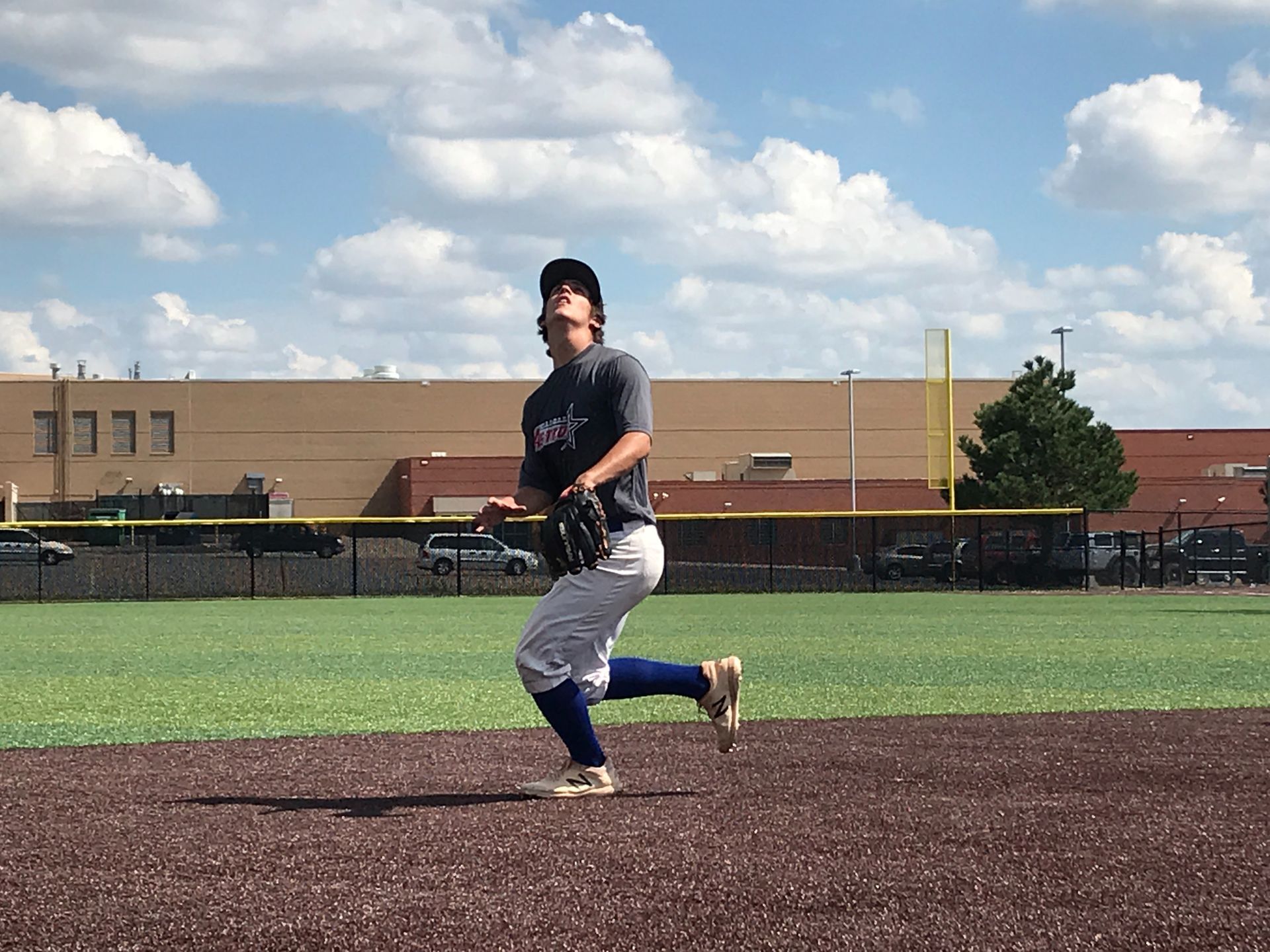 Baseball pitcher mid-throw on a dirt field under a blue sky with clouds