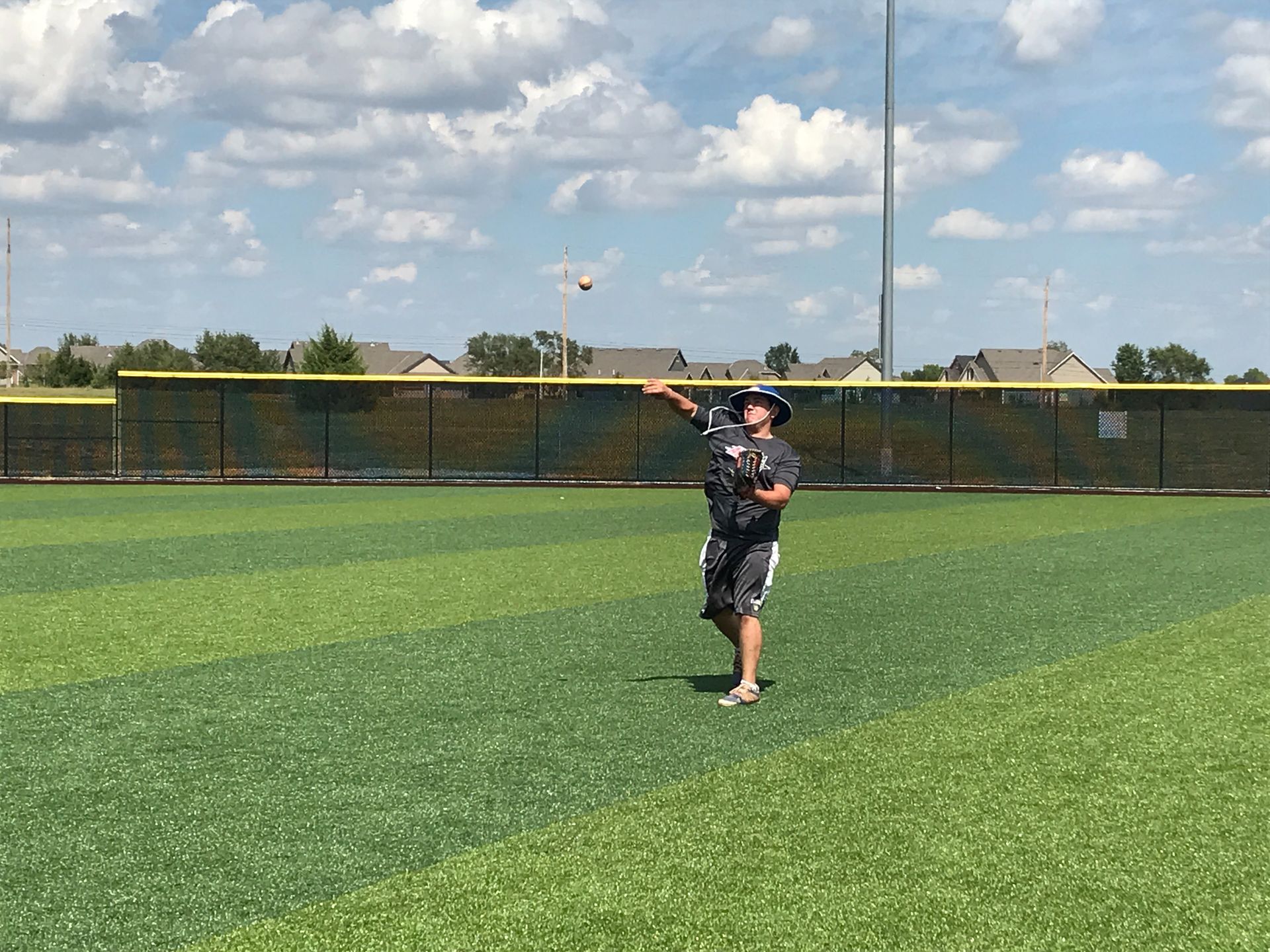 Child standing on a baseball field under a blue sky, holding a glove near the outfield fence.