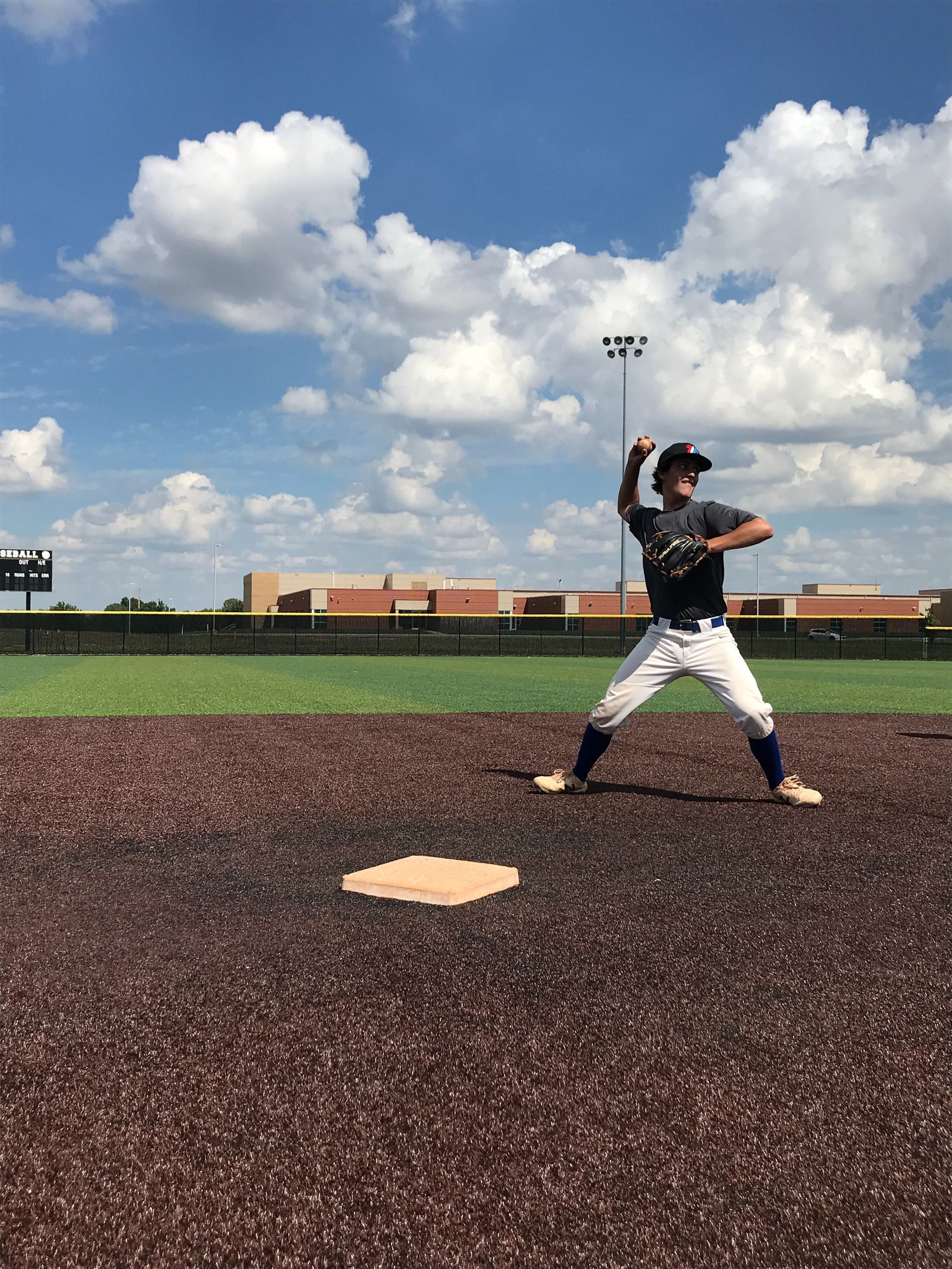 Baseball pitcher mid-throw on a field under a cloudy blue sky.