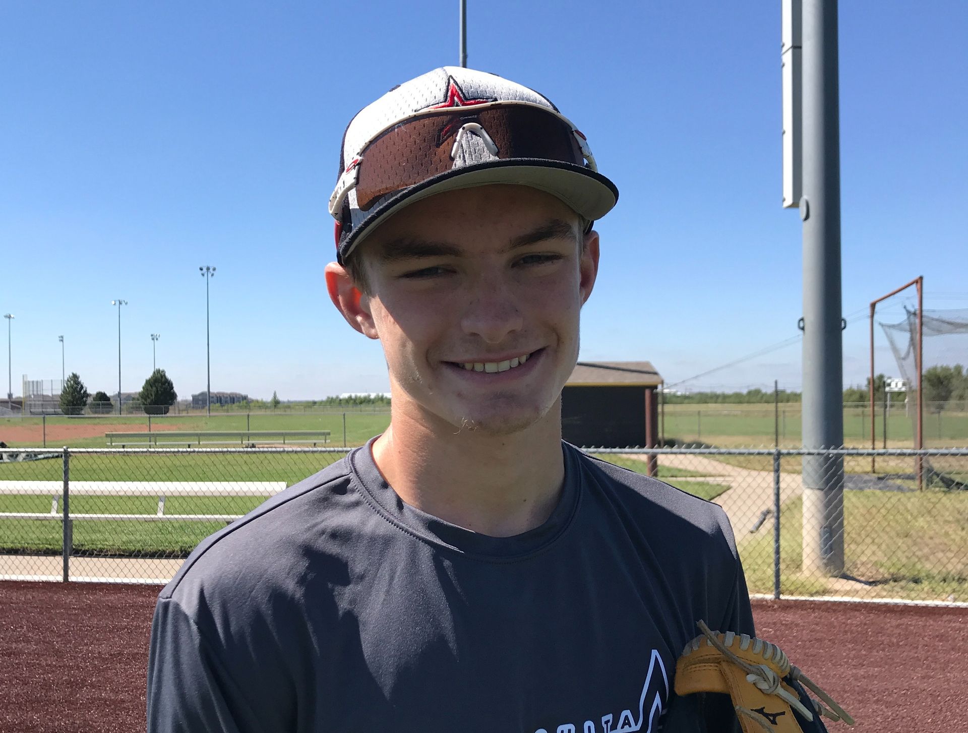 Young baseball player in a cap smiling on a field, holding a glove under a clear blue sky