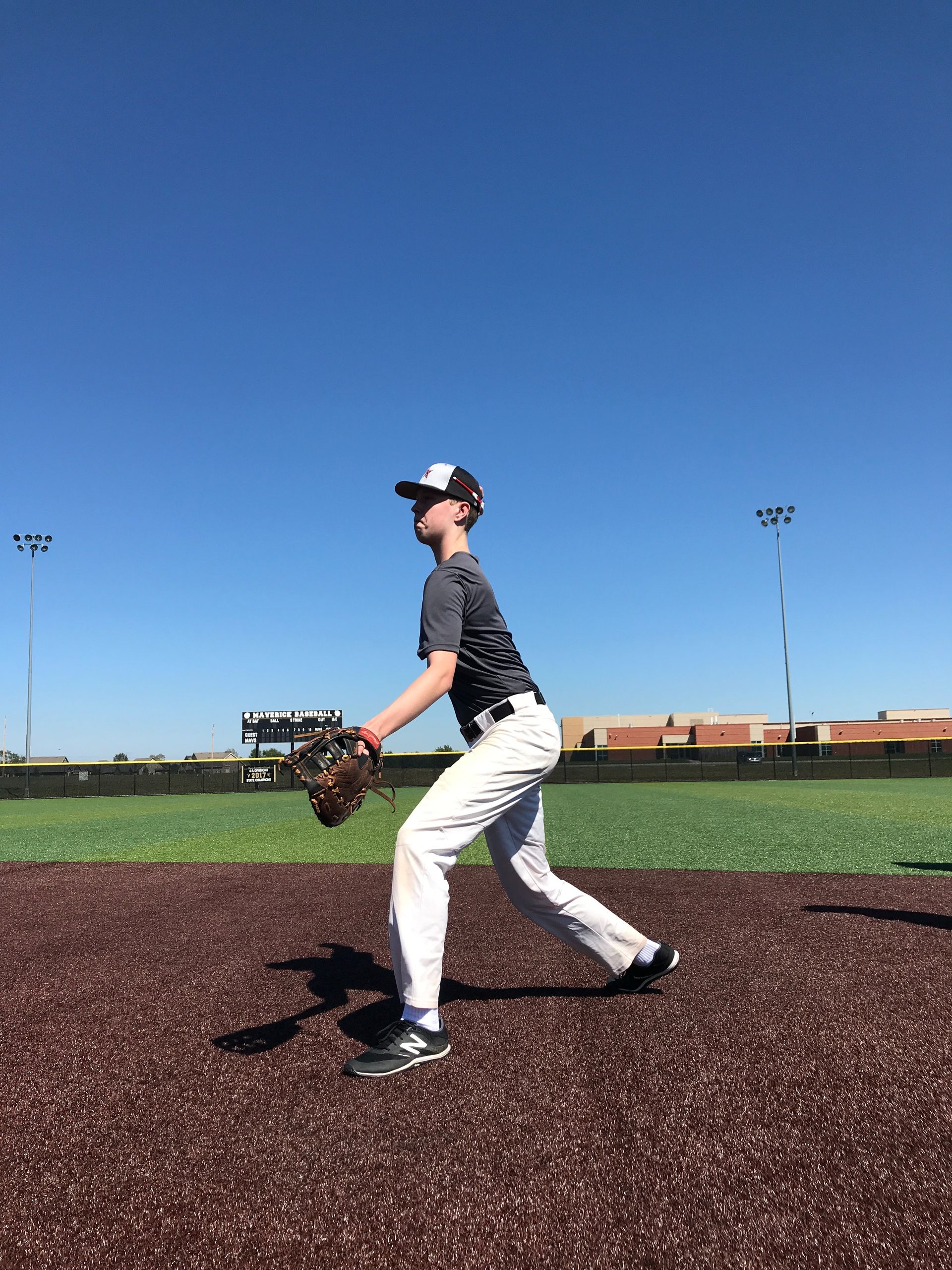 Baseball pitcher in white pants and dark jersey on the mound, holding a glove under a clear blue sky.