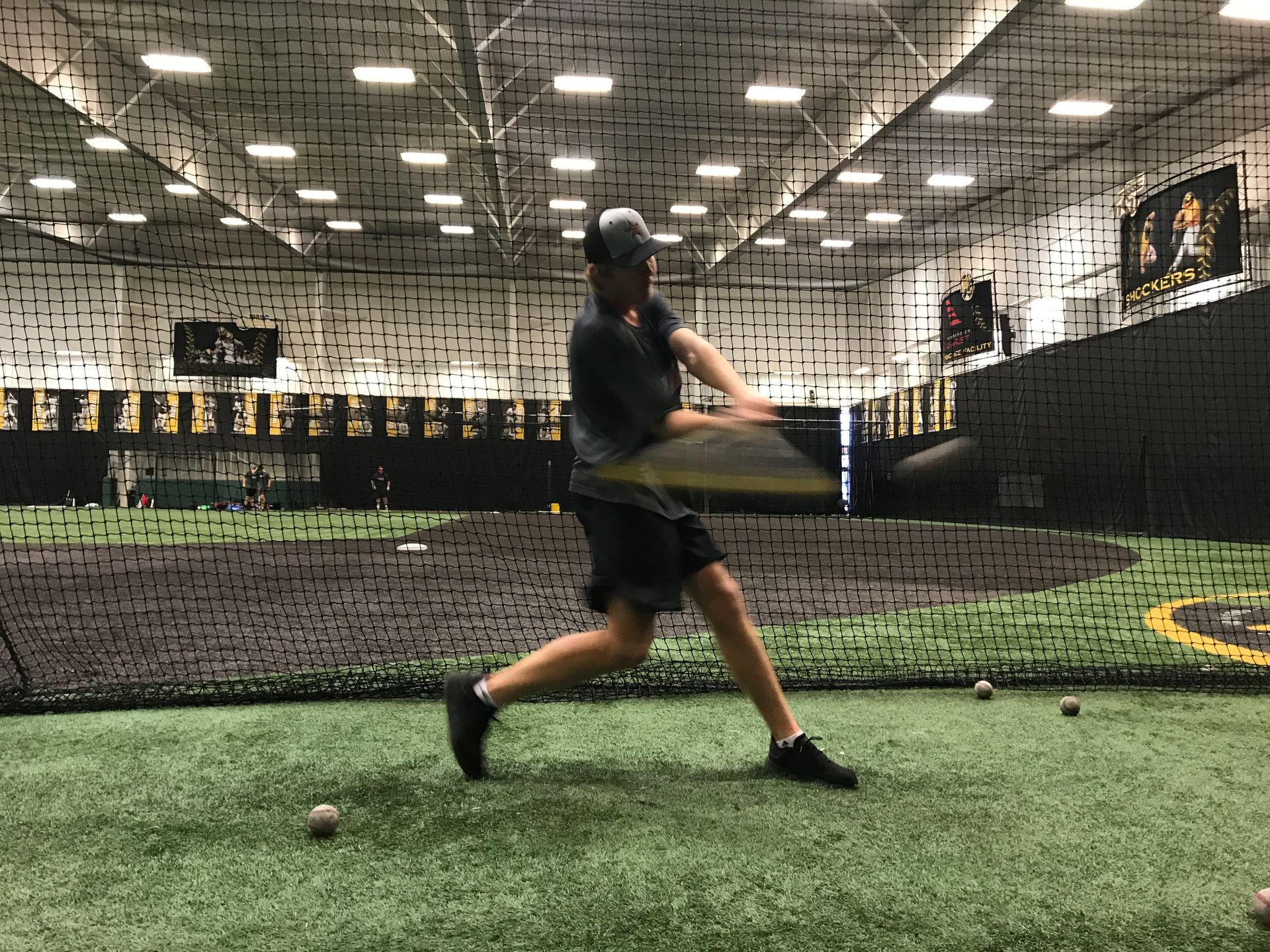 Baseball player swings bat in an indoor training cage on artificial turf.