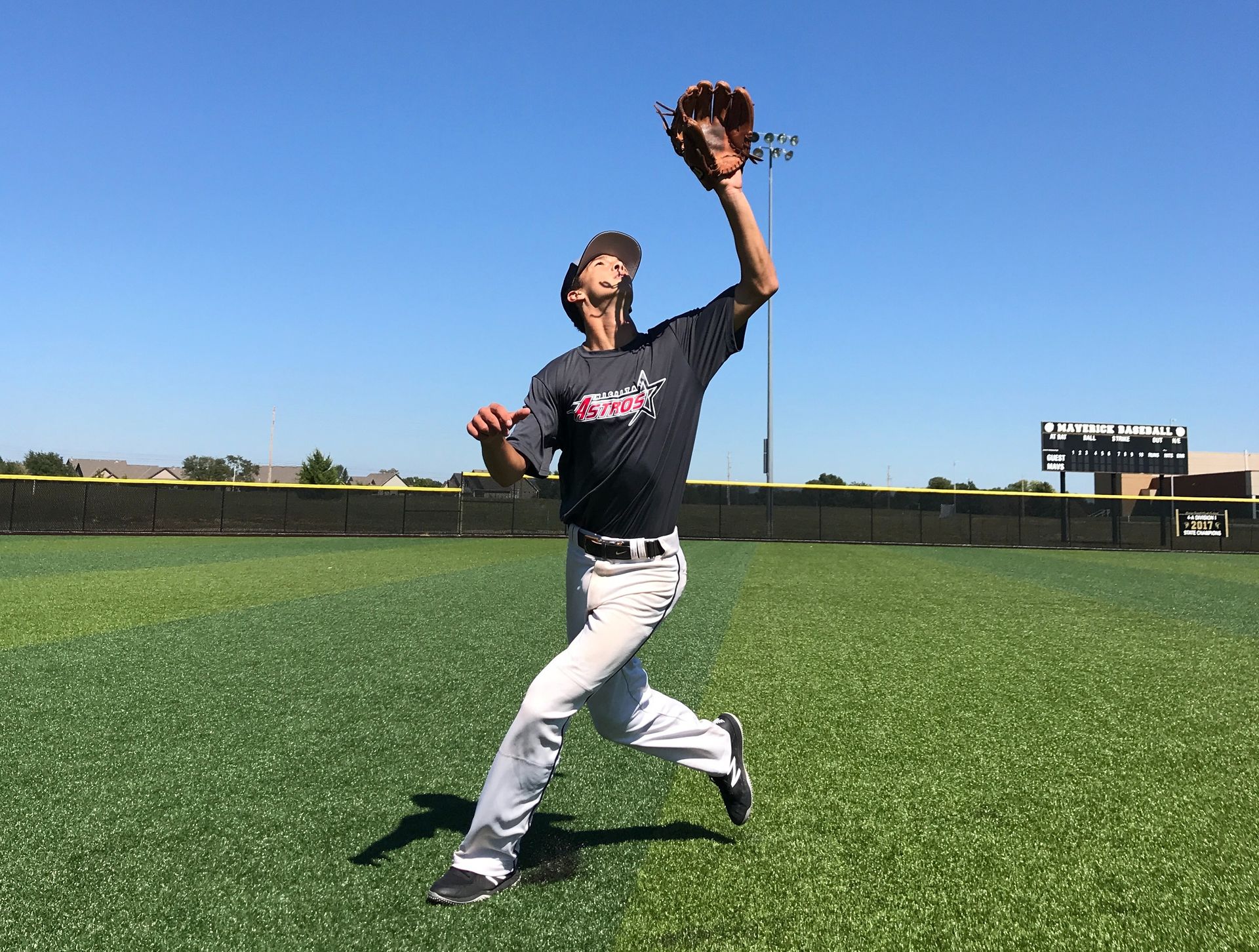 Baseball player leaping to catch a ball on a sunny field, glove raised overhead.