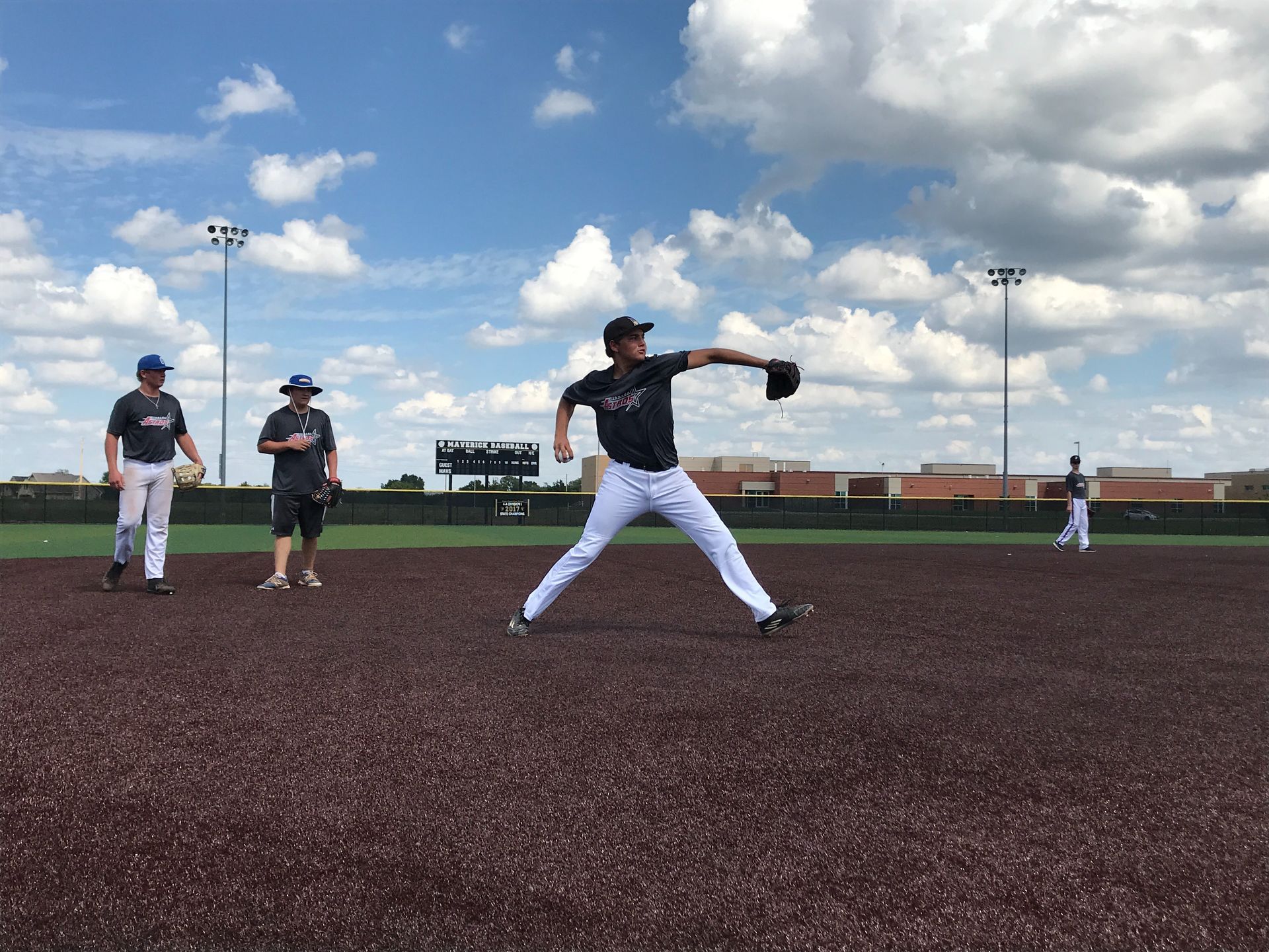 Baseball players warming up on a field under a cloudy sky, one pitcher mid-throw in the foreground.