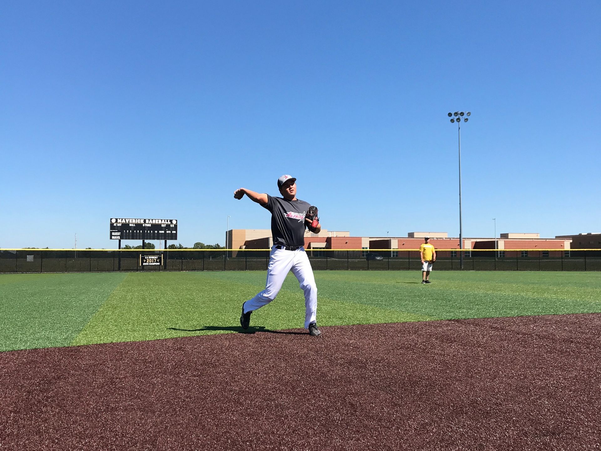 Baseball pitcher in white pants and dark jersey mid-throw on a field under a clear blue sky.