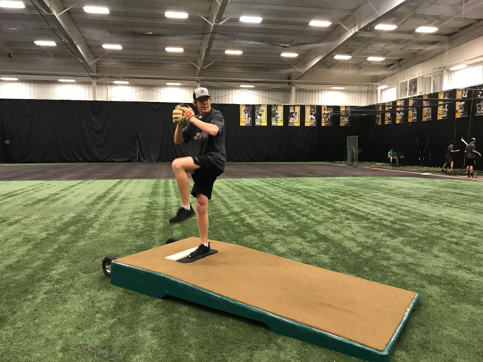 Baseball pitcher in black uniform lifts leg on a mound inside an indoor training facility.