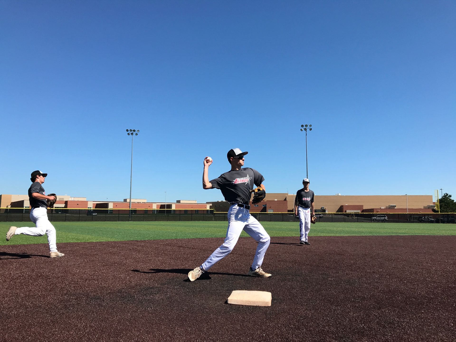 Baseball players practicing on a field, with a pitcher mid-throw near first base under a clear blue sky