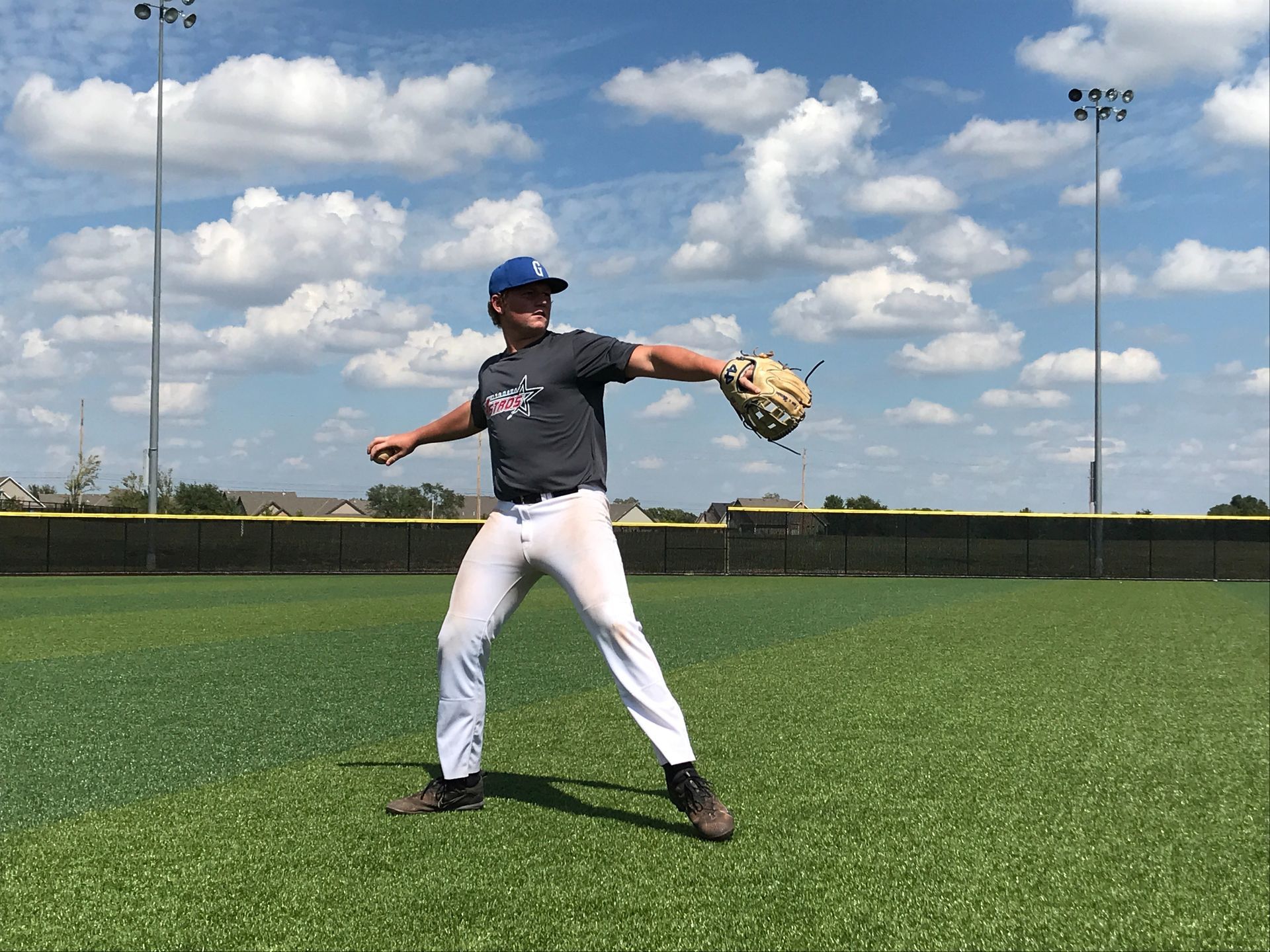 Baseball player in gray shirt and white pants throwing on a grassy field under a cloudy sky