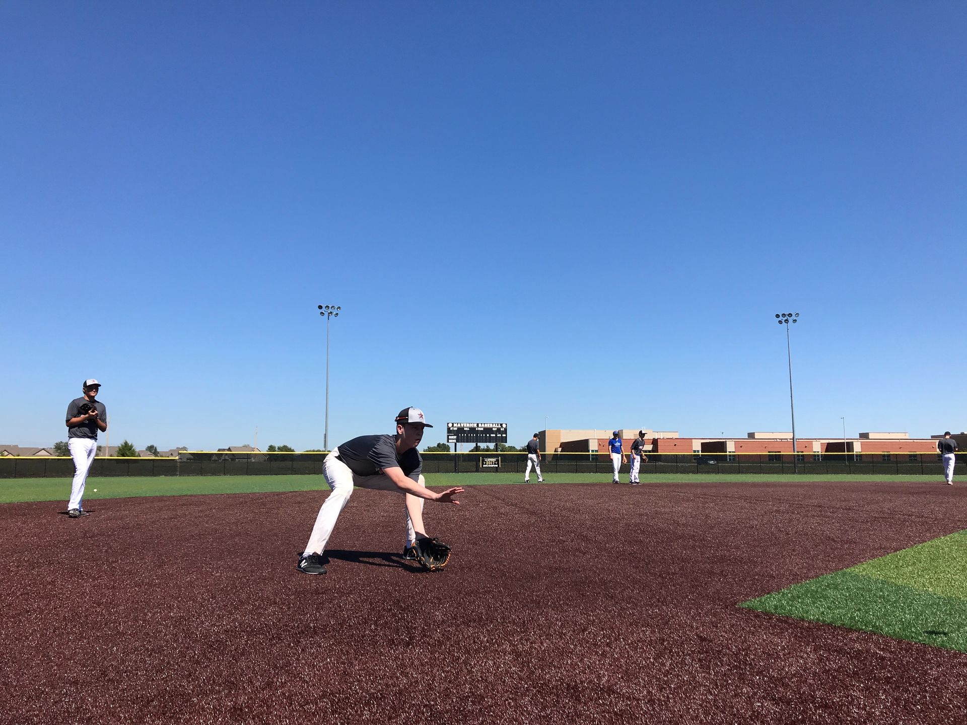 Baseball players on a field under a clear blue sky, one crouched to field a ground ball
