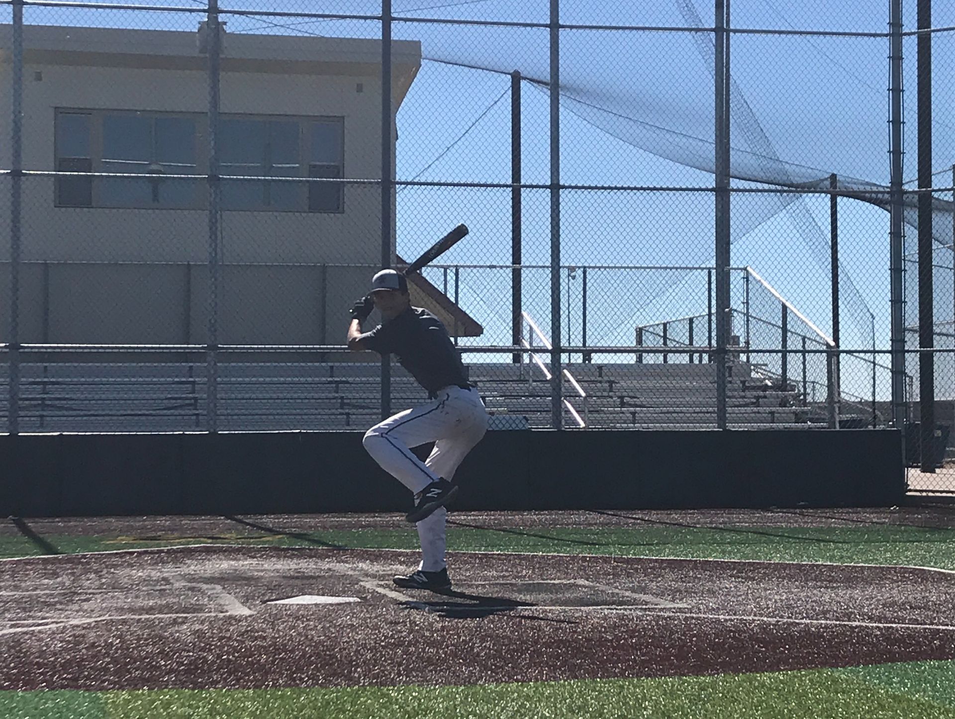 Baseball player in black jersey and gray pants prepares to swing at a batting cage.