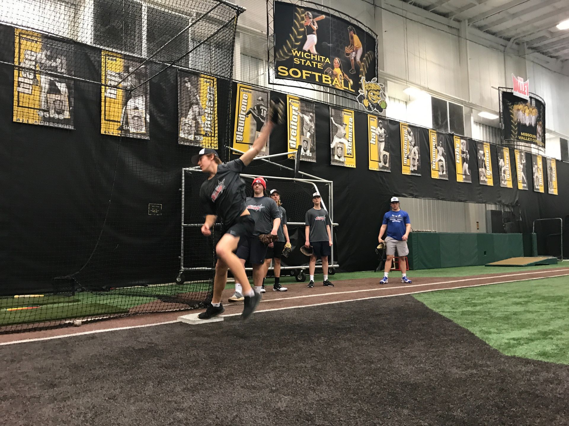 Athletes running indoor track drills on turf beside a netted wall and banners
