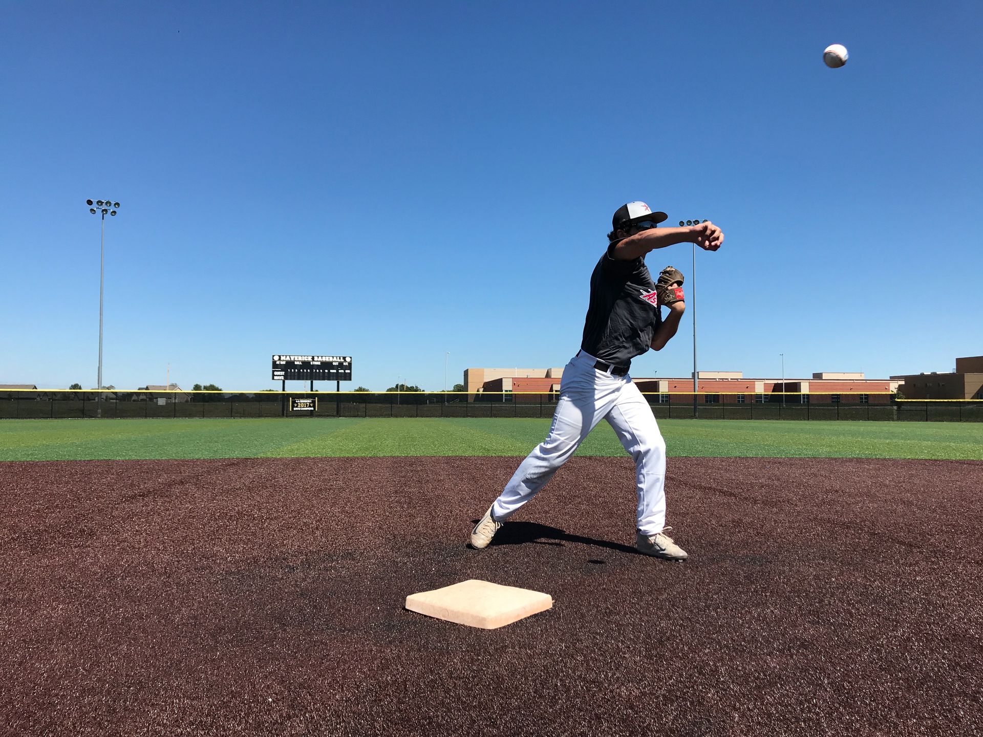 Baseball player mid-throw on a field under a clear blue sky, with a ball in the air above.
