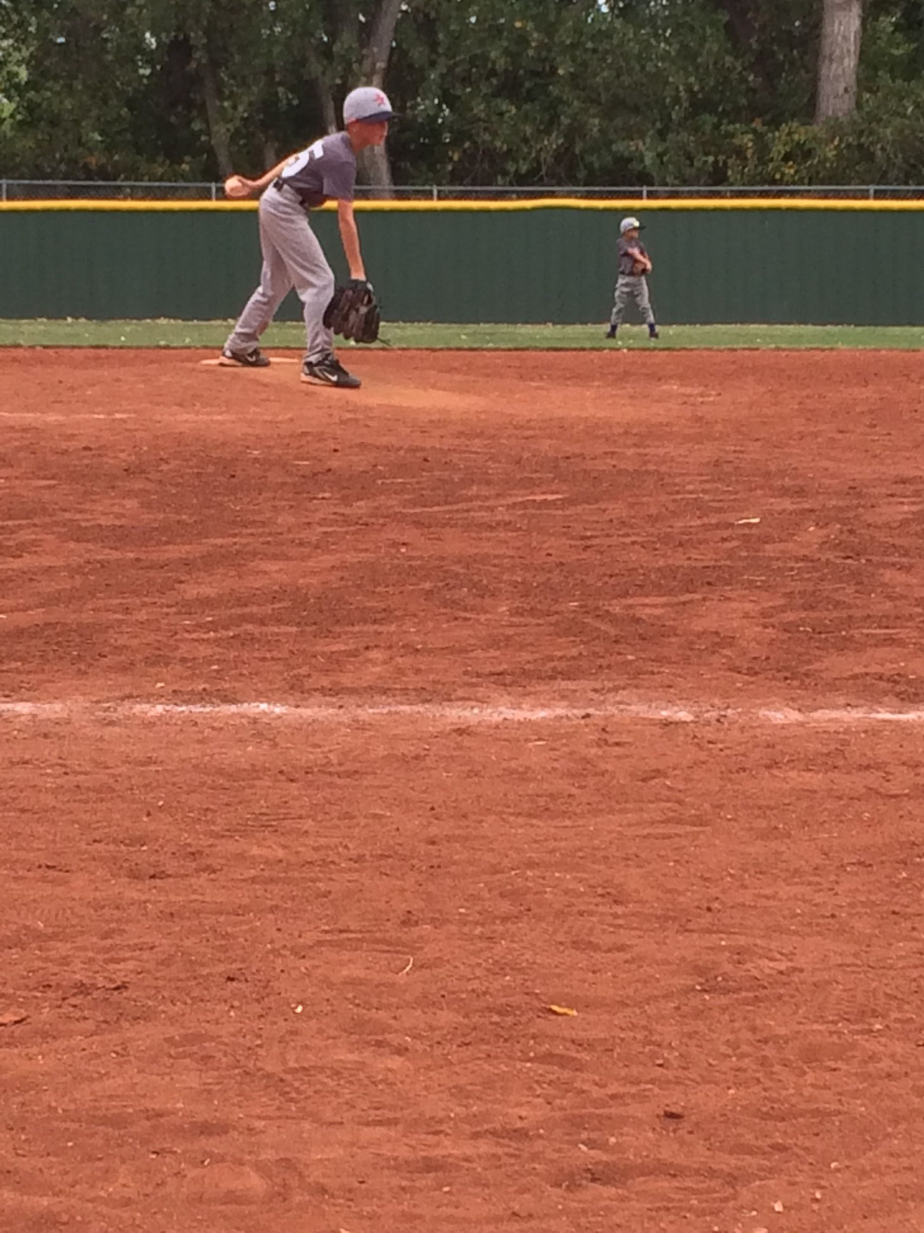 Two baseball players on a red dirt field near a green outfield fence, one readying to field a ball.