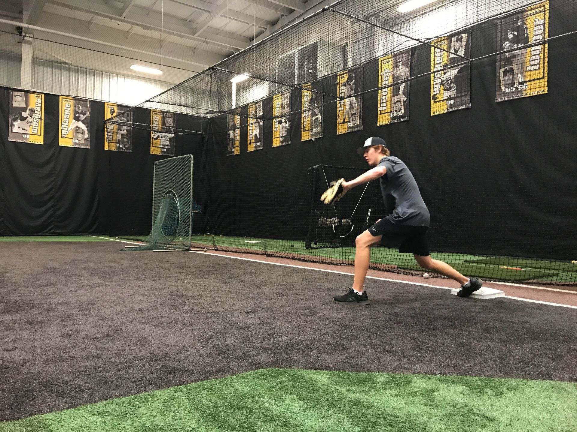 Baseball player in black swings in an indoor batting cage with black nets and turf floor