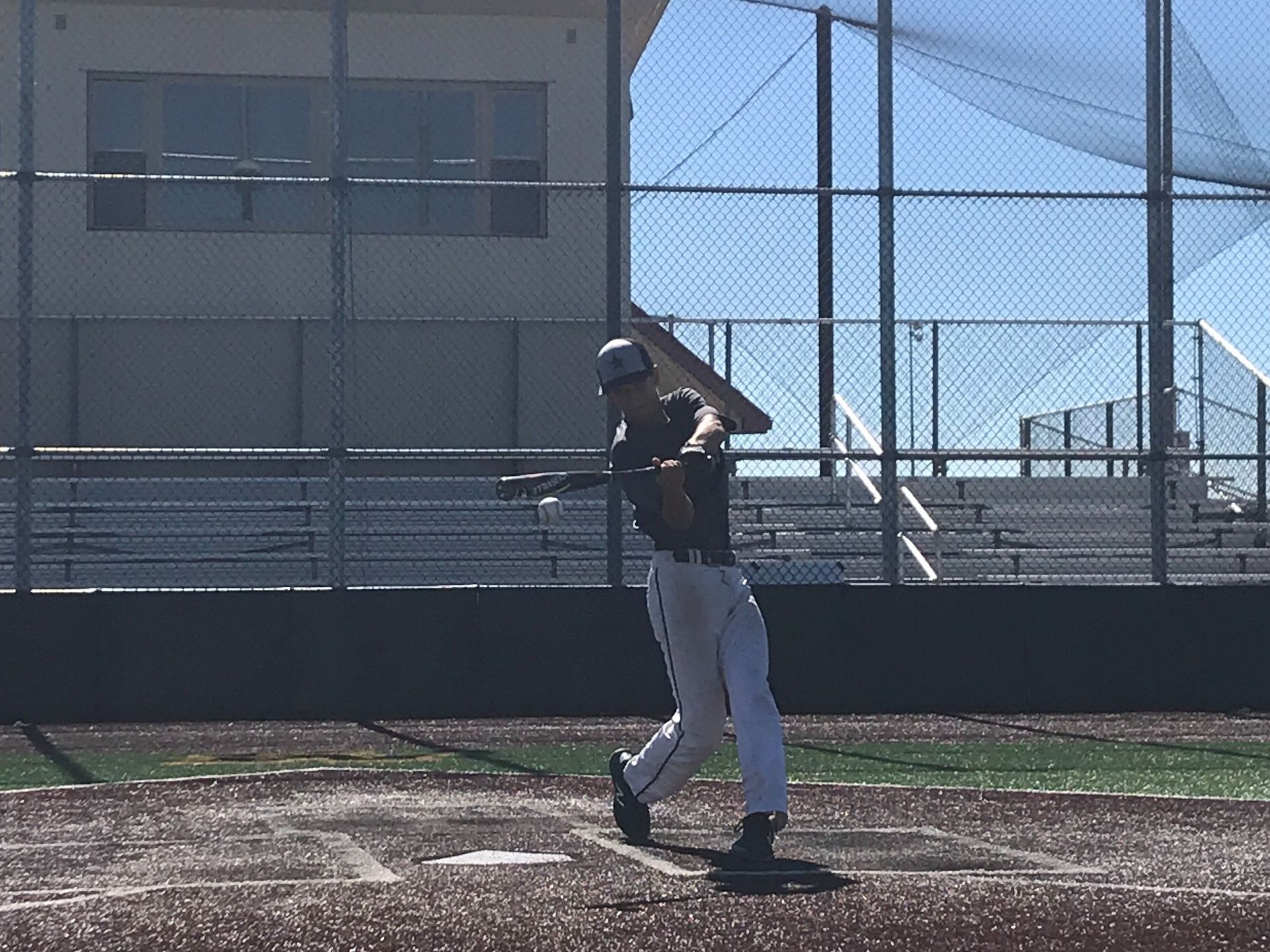 Baseball batter in a dark shirt and gray pants swinging on a dirt field near a chain-link fence.