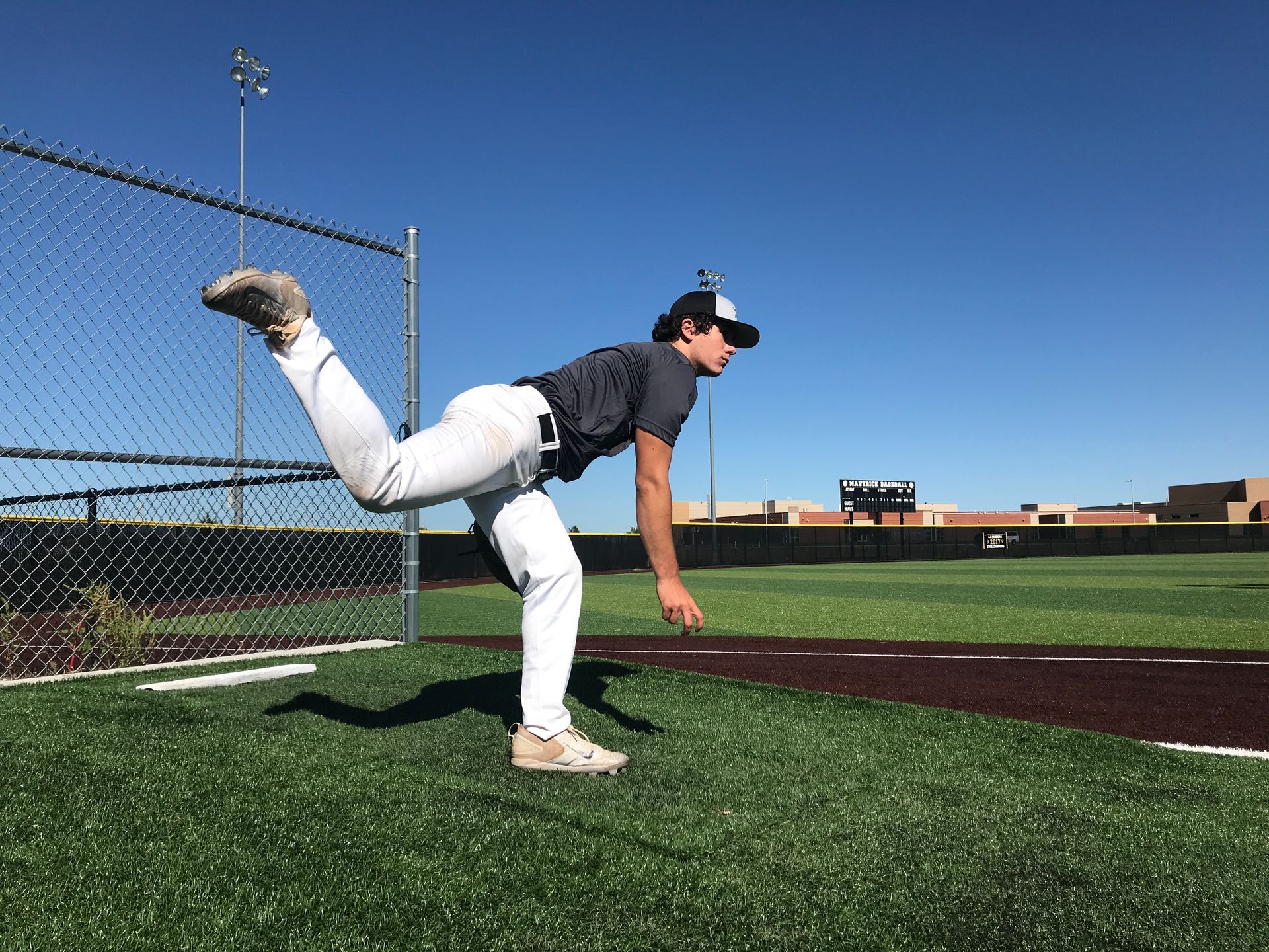 Baseball pitcher mid-throw on a sunny field, wearing white pants and a dark shirt beside a chain-link fence