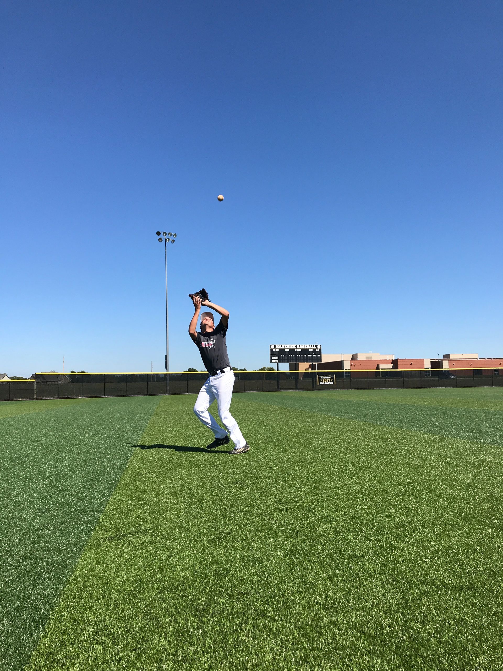 Person in white pants catches a baseball under a clear blue sky on a grassy field.