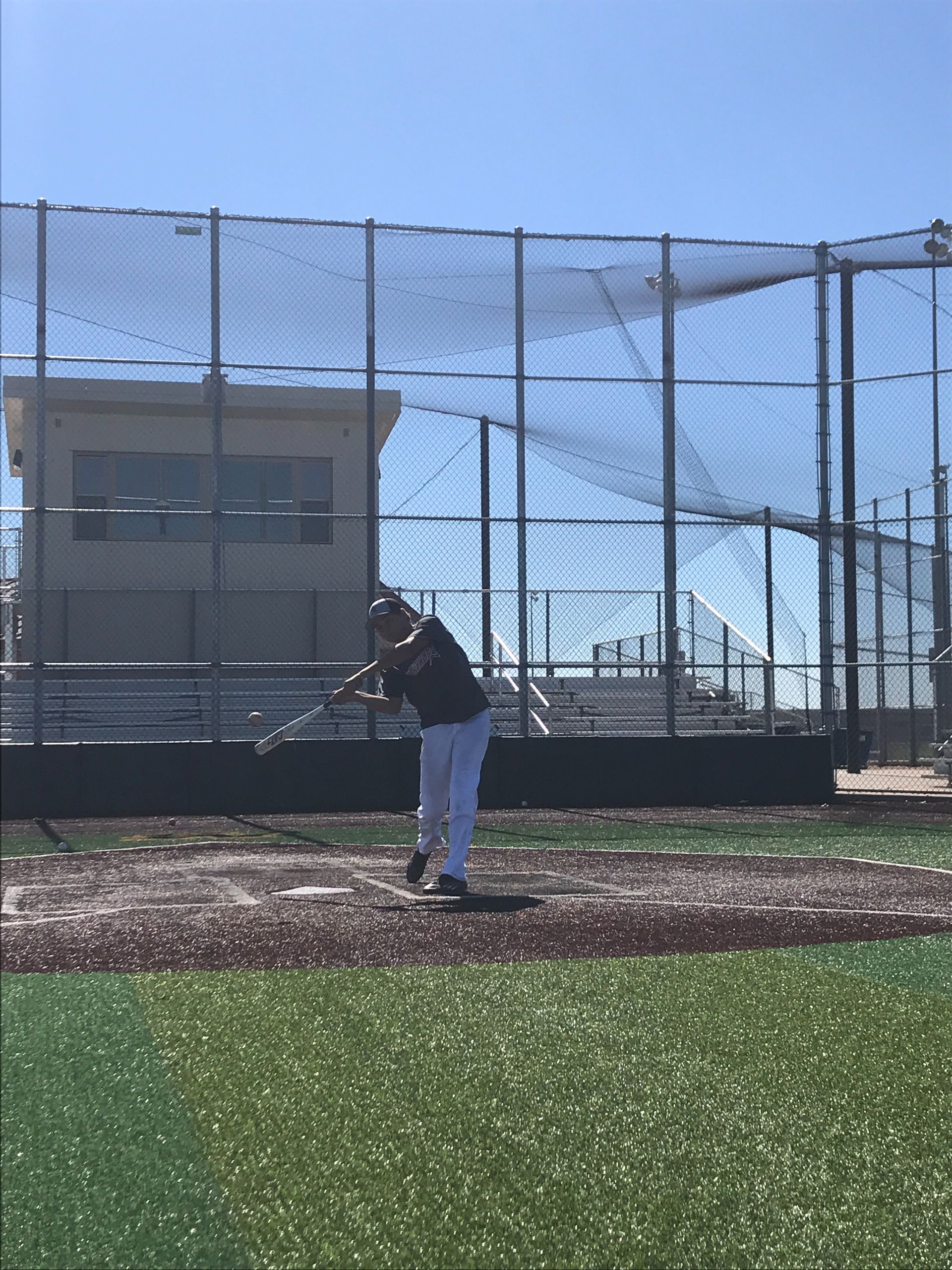 Baseball batter at home plate on a field, with a chain-link fence and building in the background.