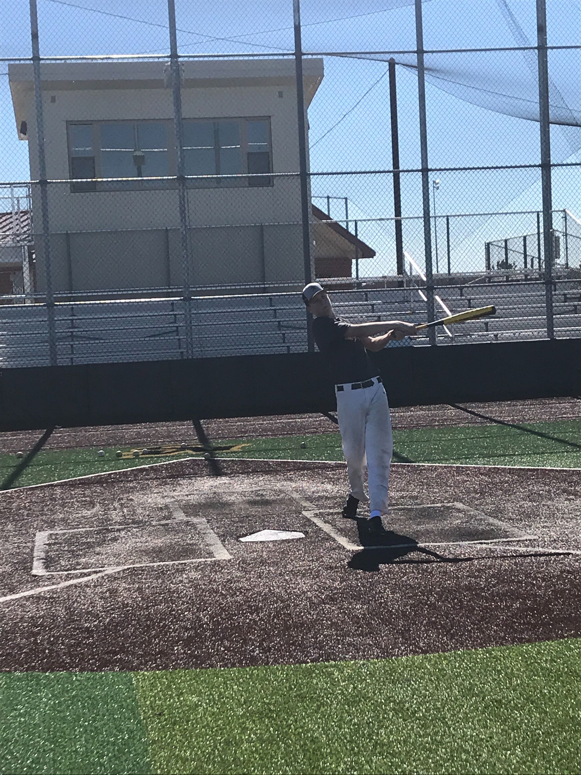 Baseball player standing on field near home plate in front of a chain-link fence and building.