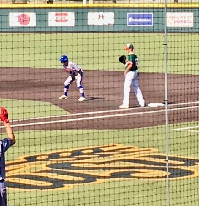Baseball runner on first base with fielder nearby, viewed through netting at a field.