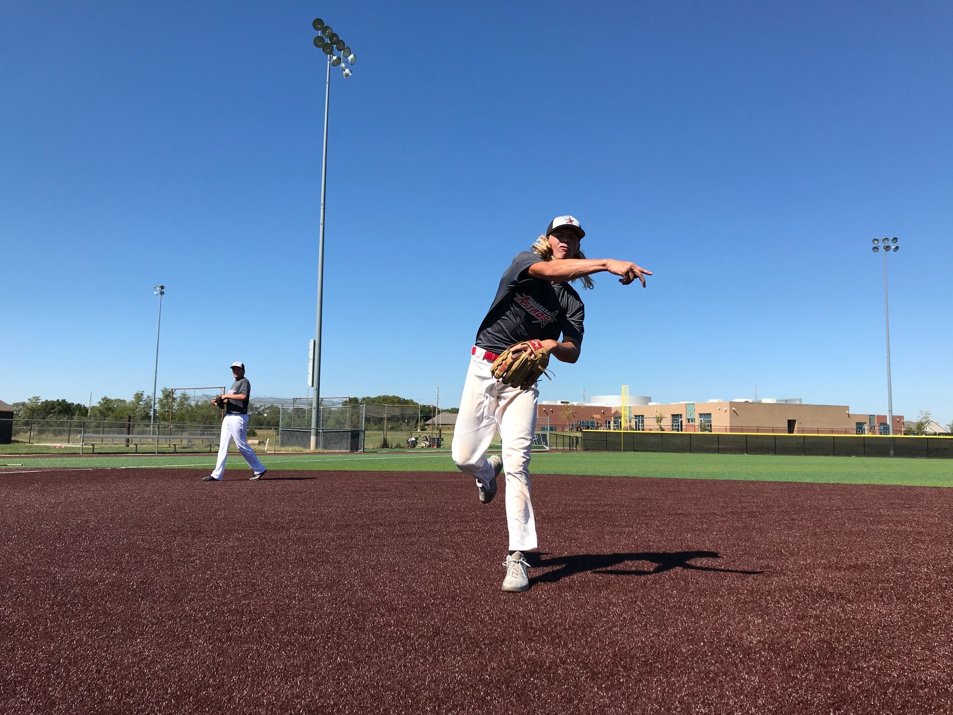 Baseball pitcher in mid-throw on a field, with another player in the background under a clear blue sky