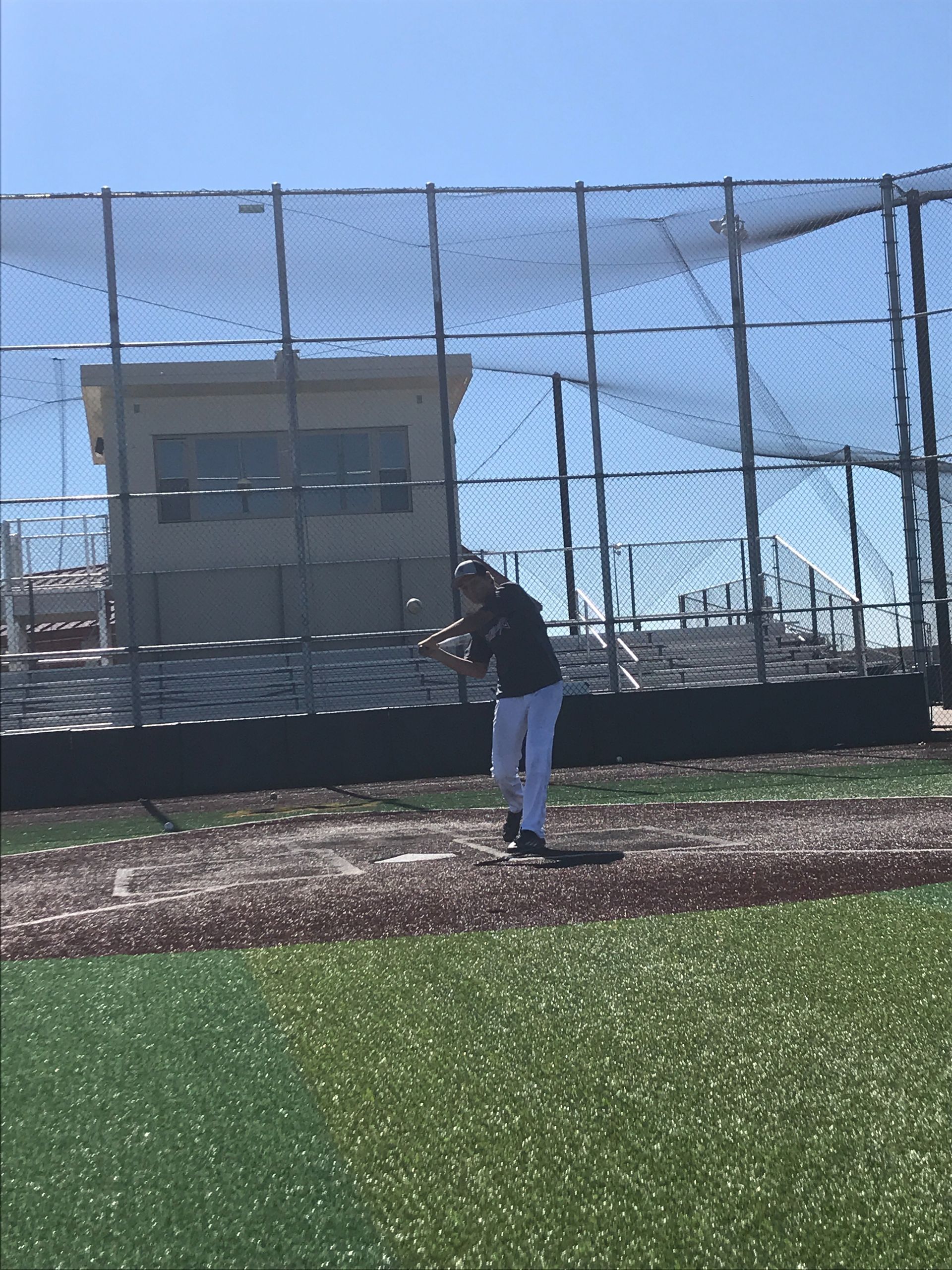 Person standing on a baseball field near the fence, holding a bat under a clear blue sky.