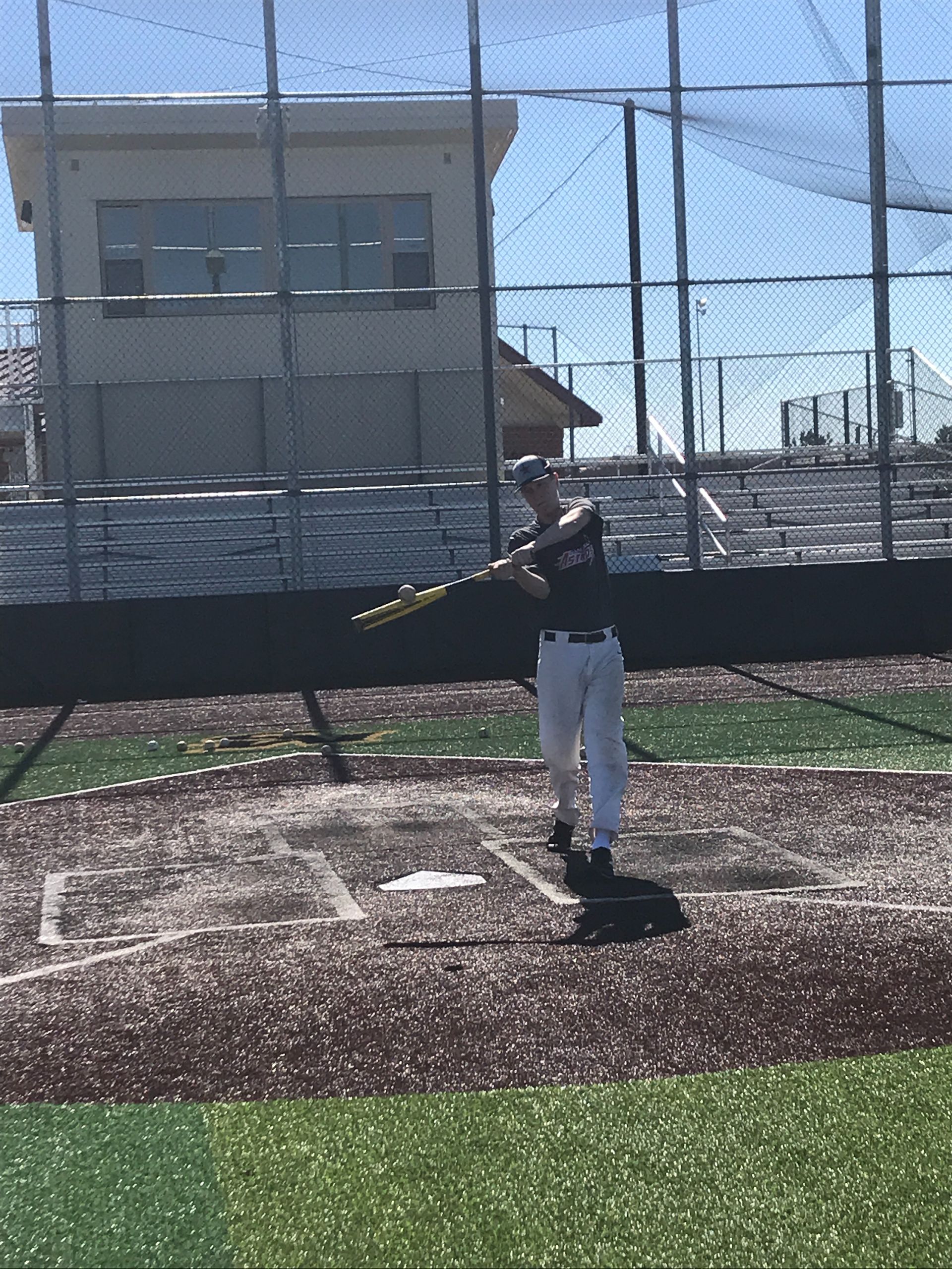 Baseball player swinging a bat on a field near home plate, with nets and a building in the background