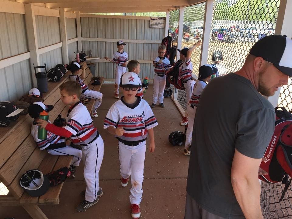 Youth baseball players in white uniforms getting ready in a dugout, with bats, helmets, and gear on benches