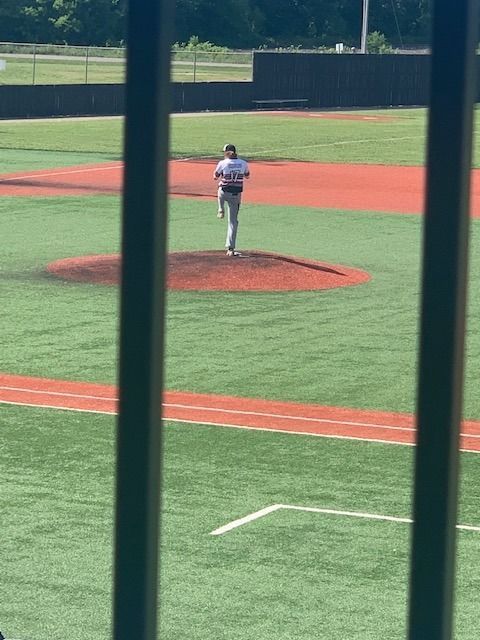 Baseball pitcher standing on the mound in a sunlit field, viewed through stadium railing.