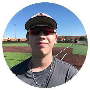 Person in sunglasses and a cap standing on a baseball field under a blue sky.