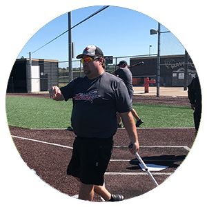 Coach on a baseball field gesturing while holding a bat, with players in the background under a clear blue sky