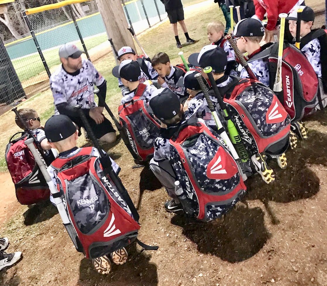 Kids with baseball gear and red-black rolling bags gathered on a dirt field near a fence.