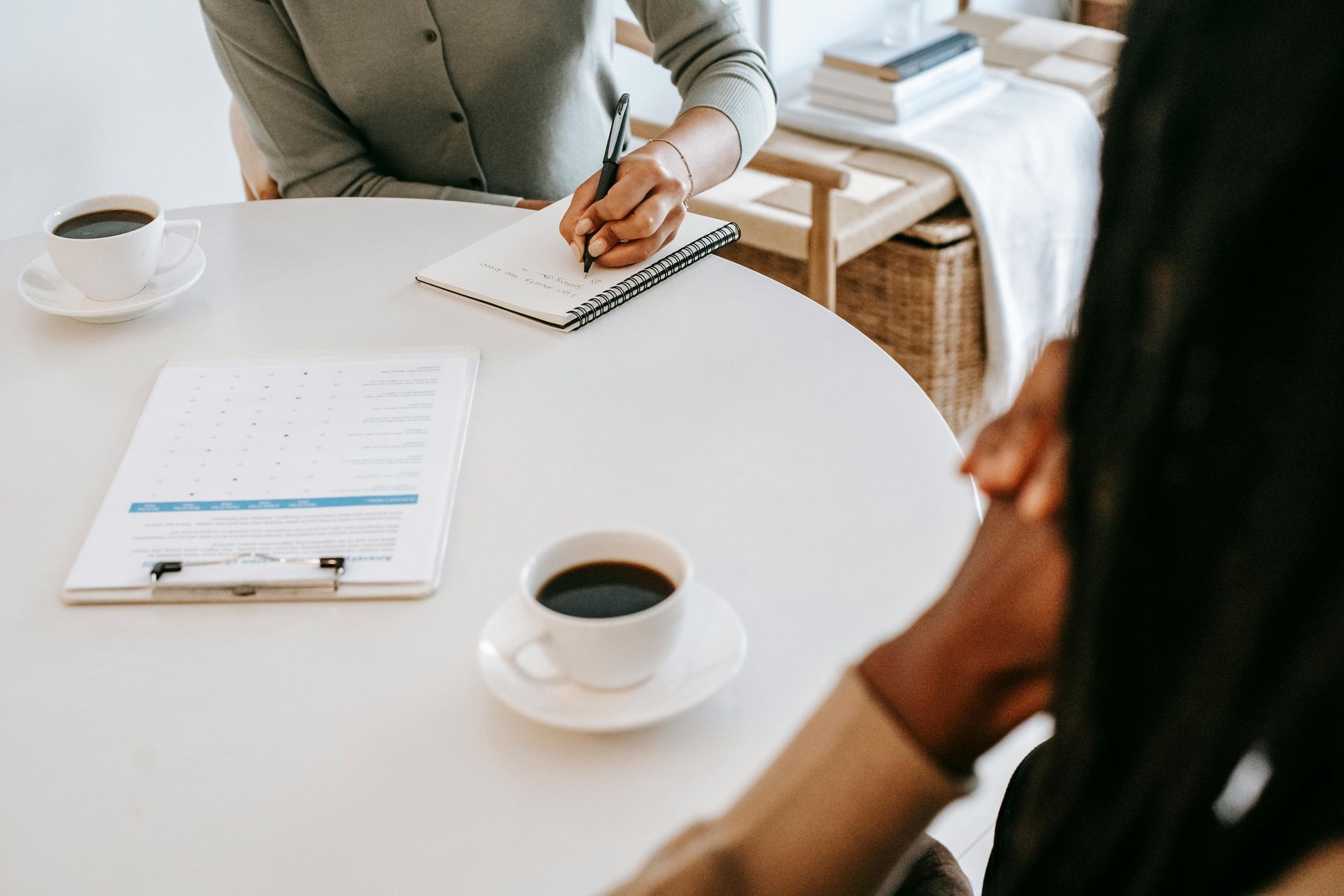 A woman is writing in a notebook while sitting at a table with a cup of coffee.