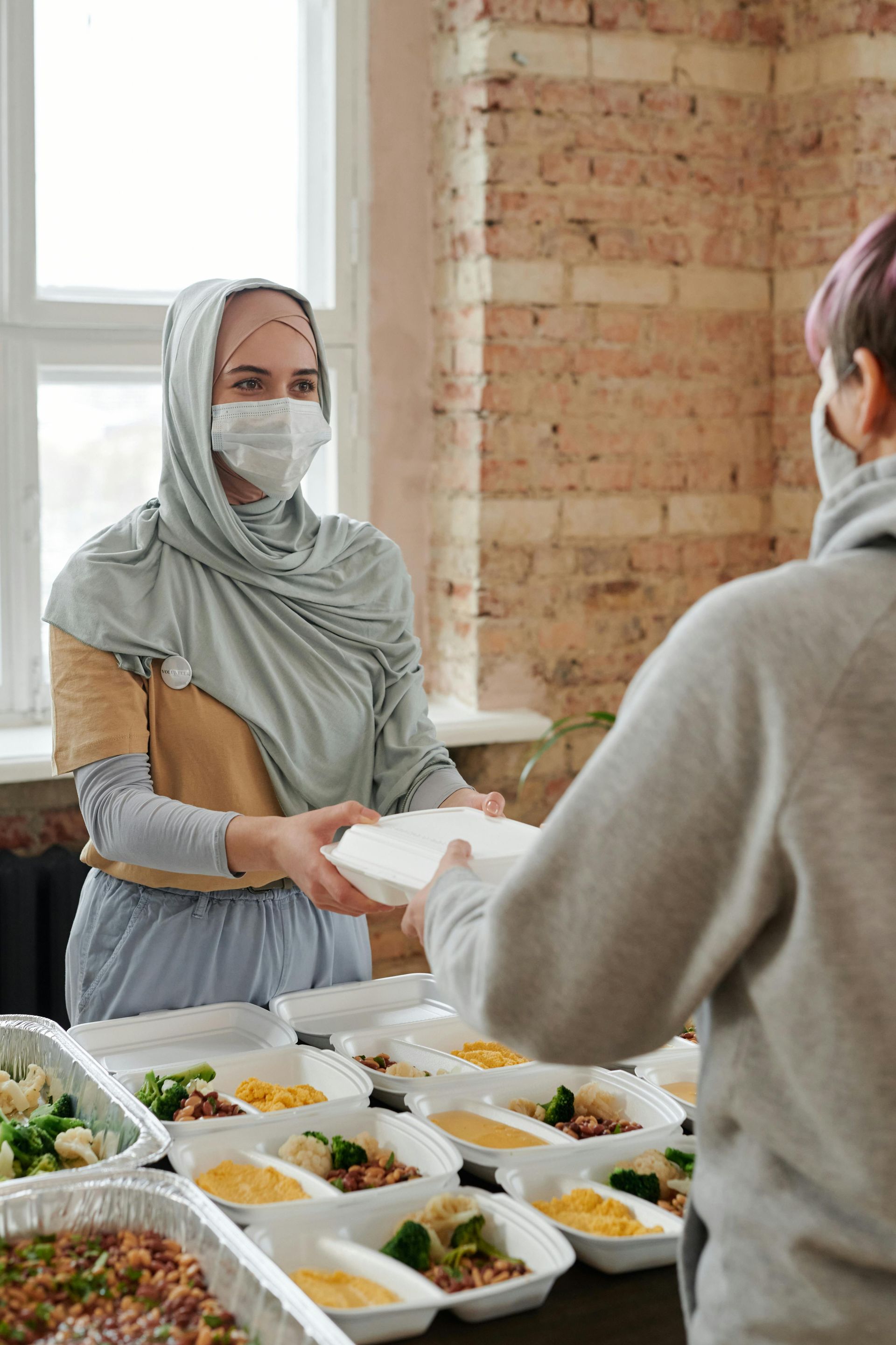 A woman wearing a mask is giving a plate of food to another woman.