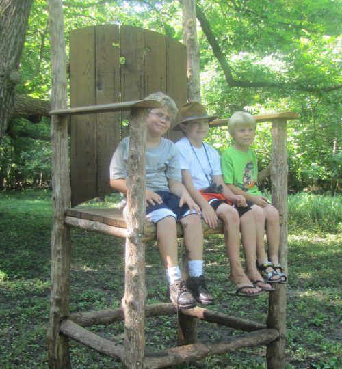 Three children are sitting on a wooden chair in the woods