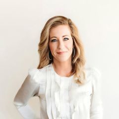 A woman wearing a white shirt and earrings is standing in front of a white wall.