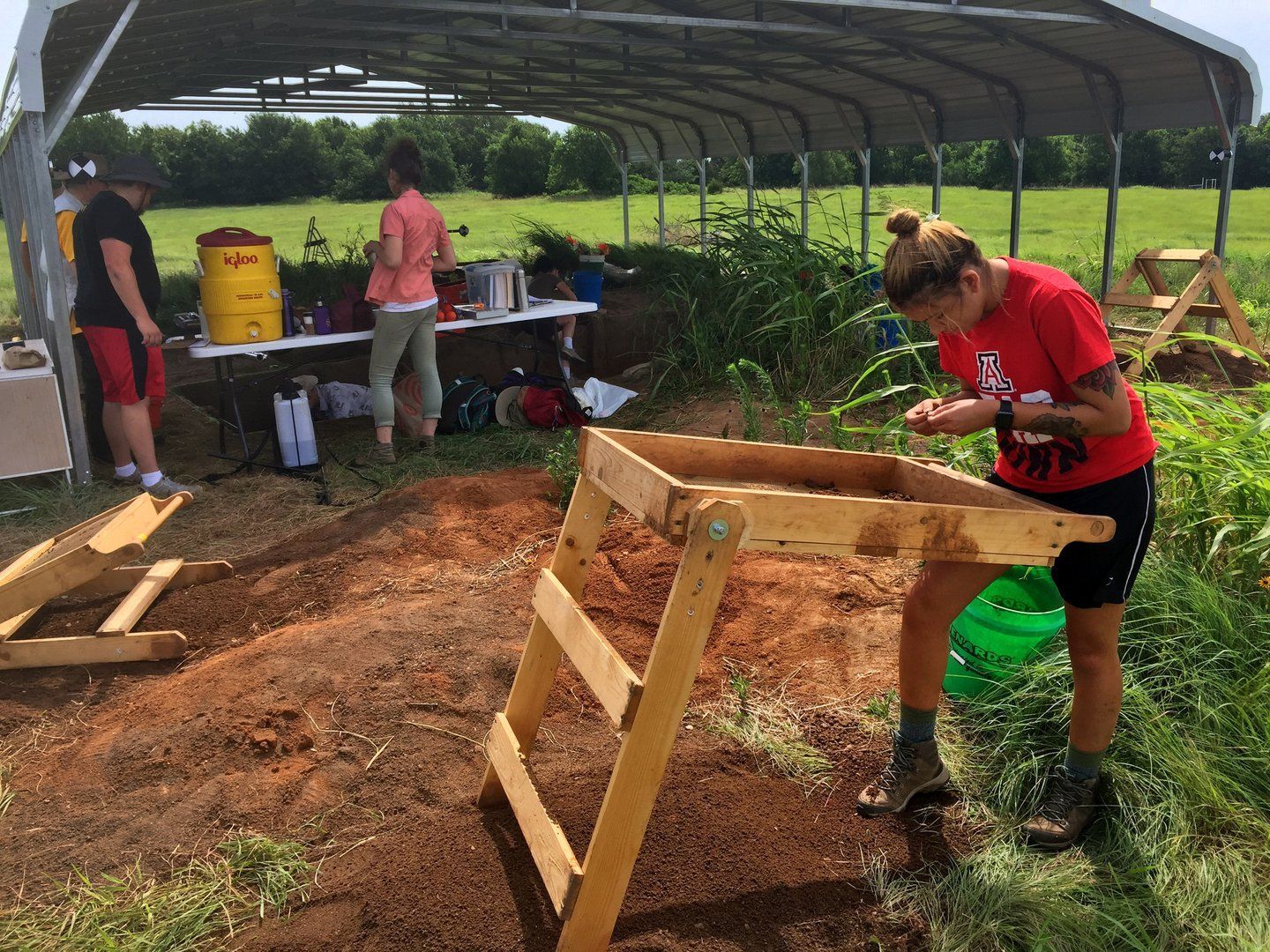 A woman in a red shirt is working on a wooden stand at an archeological dig site.