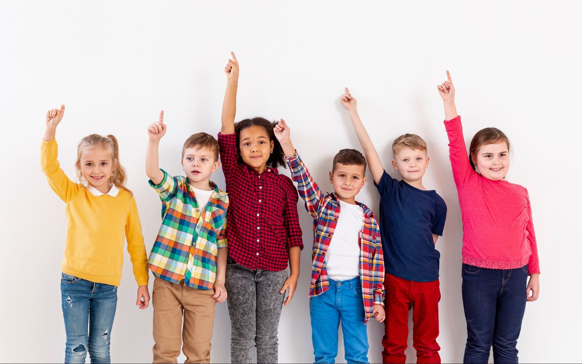 Six children standing in a row, all pointing upwards with smiles, against a white background.