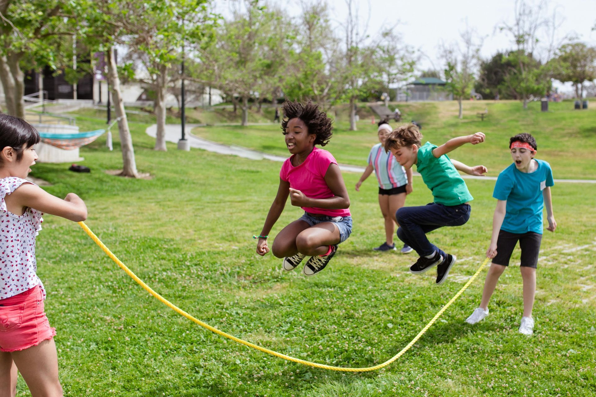 Children jumping rope in a grassy park. One girl turns the yellow rope while others jump over it.