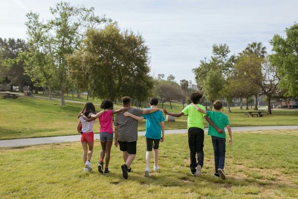 Six children with arms around each other walking on a green lawn in a park.