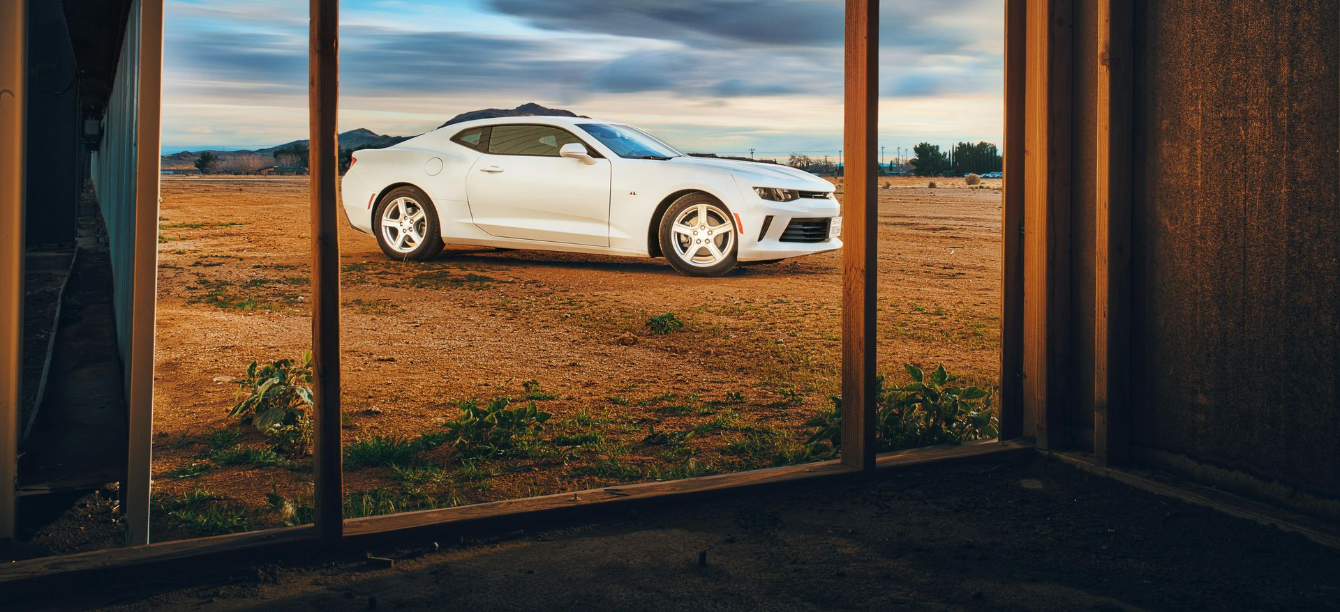 White Camaro car parked on dry land, viewed through a wooden doorway. Cloudy sky in the background.