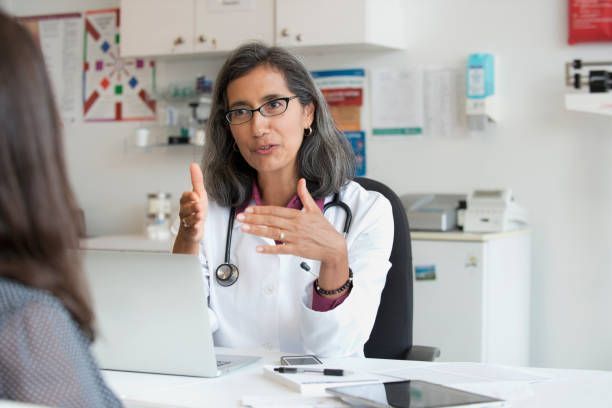 A doctor is talking to a patient while sitting at a desk in front of a laptop.