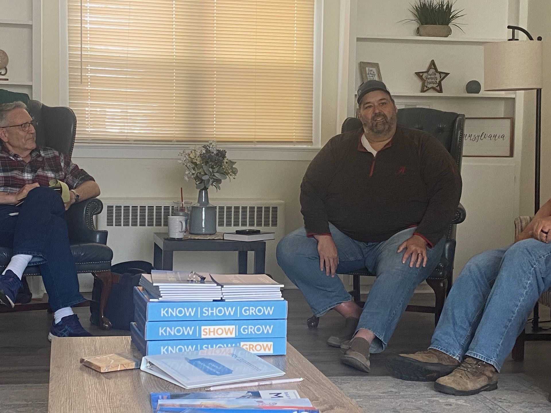 A group of men are sitting in a living room with a stack of books that say know work grow
