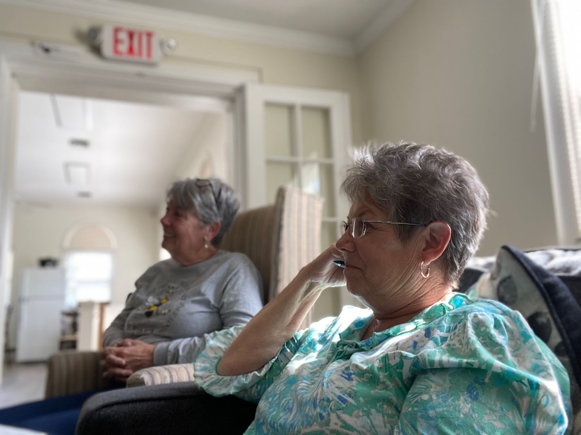 Two older women are sitting in chairs in front of an exit sign.
