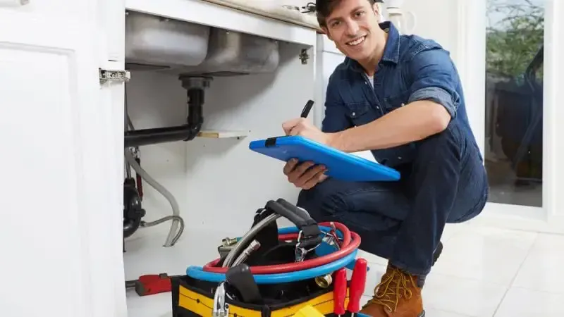 A plumber is kneeling down under a sink and writing on a clipboard.