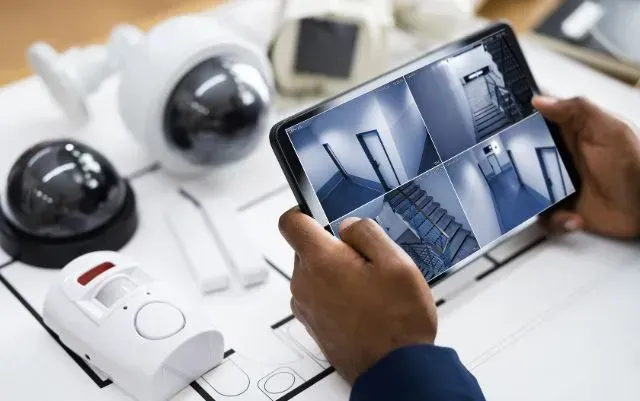 Hand holding a tablet showing security camera views, with white surveillance cameras on a desk