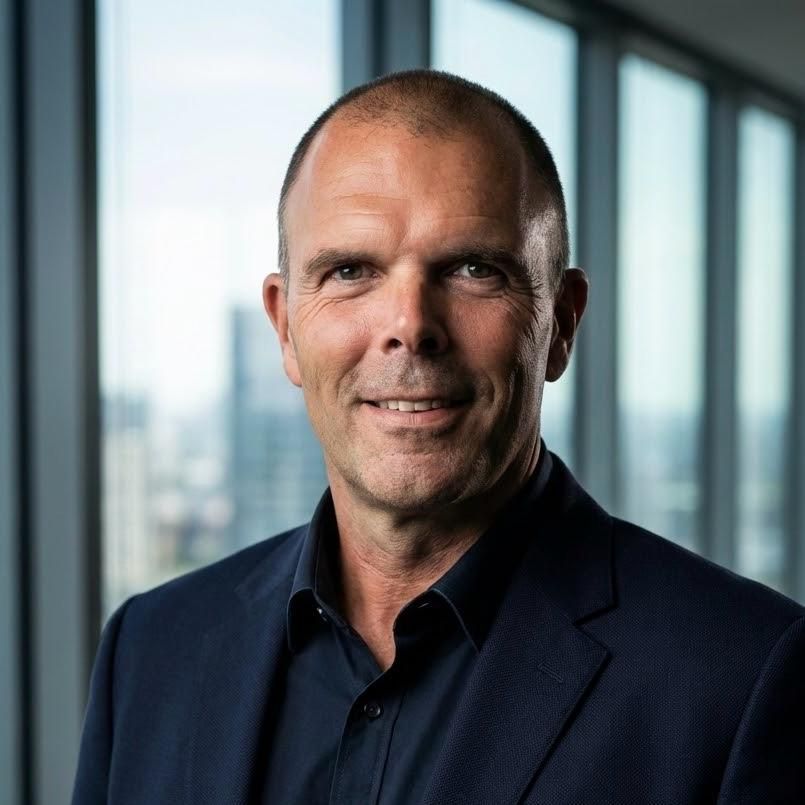 Man in dark suit smiles, posed in front of cityscape windows.
